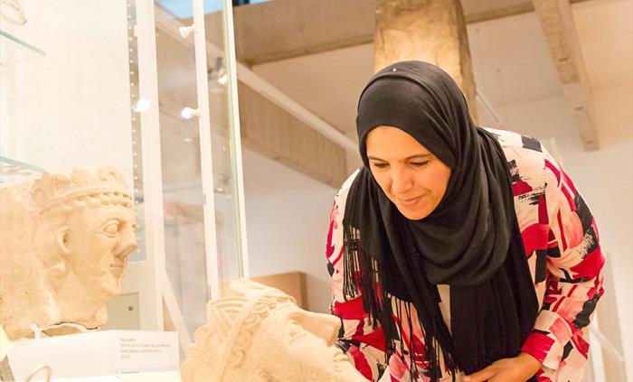 a lady wearing a black hijab and red patterned blouse is leaning forward to look at a stone carving. The carving shows the head of a medieval prince.